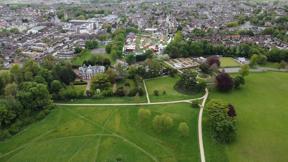 An aerial view of a large park showing well-maintained lawns, winding pathways, and various trees with light green and darker foliage. The park features open grassy areas intersected by dirt and grave