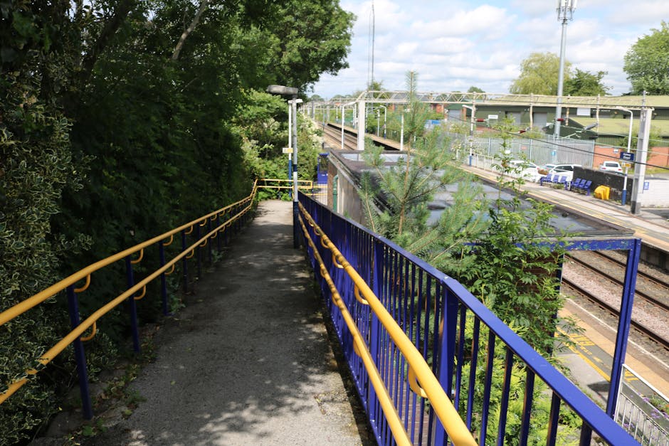 A narrow pedestrian pathway with a gravel surface, bordered by a yellow-and-blue metal safety railing, runs alongside a railway station area visible in the background. To the left of the path, dense g